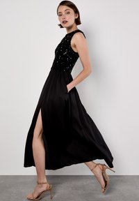 Abito da sera nero con bodice di paillettes, design senza maniche, gonna in satin fluente, spacco laterale, e abbinato a scarpe con tacco nude.