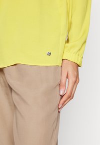 Blouse jaune à manches longues avec poignets élastiques portée avec un pantalon beige, montrant la main droite d'une personne reposant à côté d'elle.