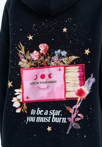 Černá látka s otevřenou krabičkou zápalek, růžové zápalky, květiny, hvězdy a text: "Zůstaň ve svém kouzle" a "Abys byl hvězdou, musíš hořet."