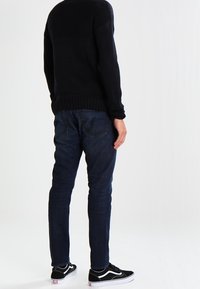 Černý pletený svetr, tmavě modré džíny a černé tenisky. Svetr má žebrovaný lem, zatímco džíny mají zadní kapsy a přiléhavý střih.