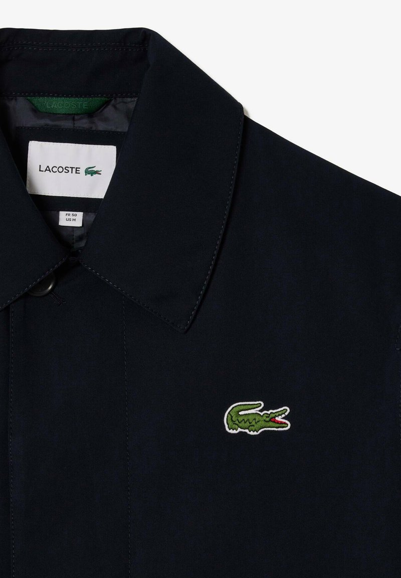 Chaqueta Lacoste de color azul marino con cuello estructurado, cierre de botones y logo de cocodrilo bordado en verde en el pecho.