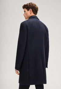 Marineblauer Wollmantel mit strukturierter Oberfläche, einreihigem Design und dezentem Karomuster. Verfügt über einen Reverskragen und knöpfbare Manschetten.