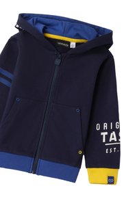 Sudadera con cremallera de color azul marino, con un dobladillo acentuado en amarillo, bolsillos frontales, forro interno azul oscuro y texto gráfico blanco en la manga.