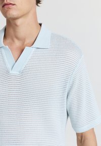 Mann trägt ein hellblaues, kurzärmliges Strick-Polohemd mit einem strukturierten, offenen Gewebe-Muster und einem Kragen vor einem schlichten Hintergrund.