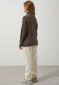 Brązowy sweter z golfem o luźnym kroju i ściągaczami, zestawiony z kremowymi spodniami oraz sneakersami, widok z tyłu.