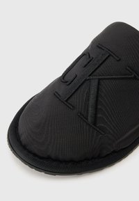 Fekete szövetpapucs kiemelkedő hímzett monogrammal, lekerekített formával, texturált felülettel és kontrasztos fekete csíkkal a talpon.