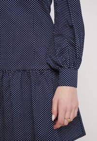 Vestido azul marino con lunares blancos, que presenta mangas abullonadas, un busto ajustado y una falda acampanada. Tejido suave con una textura sutil.