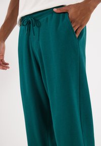 Teal sweatpants made of soft fabric with an elastic waistband, drawstring closure, and side pockets. Smooth texture with a relaxed fit.