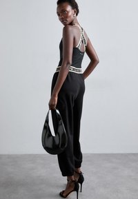 Melns bez piedurknēm tops ar kontrastējošām zīmola siksnām, apvienots ar melniem sporta biksēm ar elastīgu apakšējo daļu un spīdīgu melnu rokassomu.