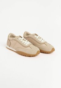 Sneakers beige con un design in materiali misti, lacci e una suola in gomma testurizzata, esposti su uno sfondo bianco.