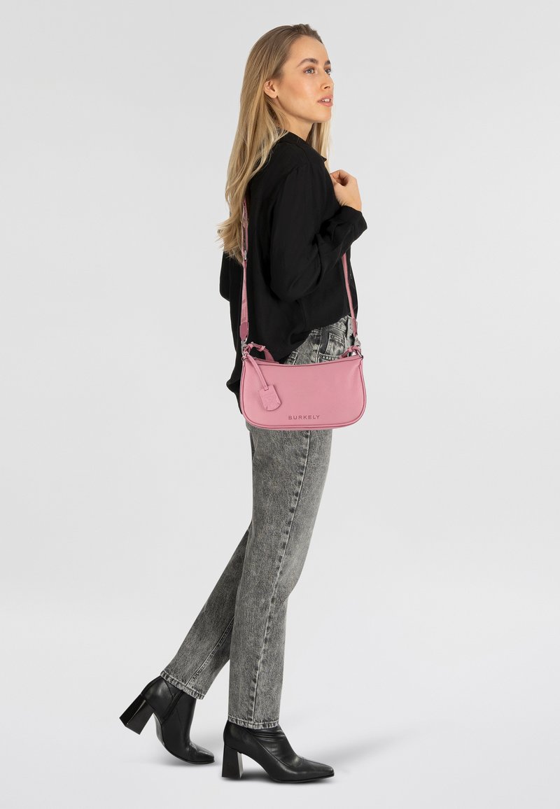 Borsa a tracolla in pelle rosa con finitura testurizzata, hardware in argento e tracolla regolabile, esposta insieme a stivali neri con tacco e jeans grigi.
