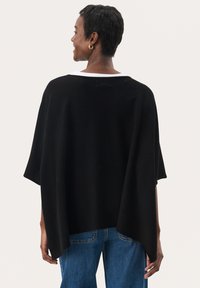 Haut style poncho noir en matériau doux, présentant des manches courtes, un col large et une coupe fluide, porté sur un jean bleu.