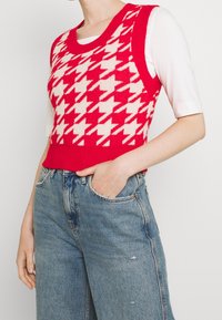 Czerwona i kremowa dzianinowa kamizelka w pepitkę z okrągłym dekoltem, noszona na białej dopasowanej bluzce i zestawiona z jasnoniebieskimi dżinsami z wysokim stanem.