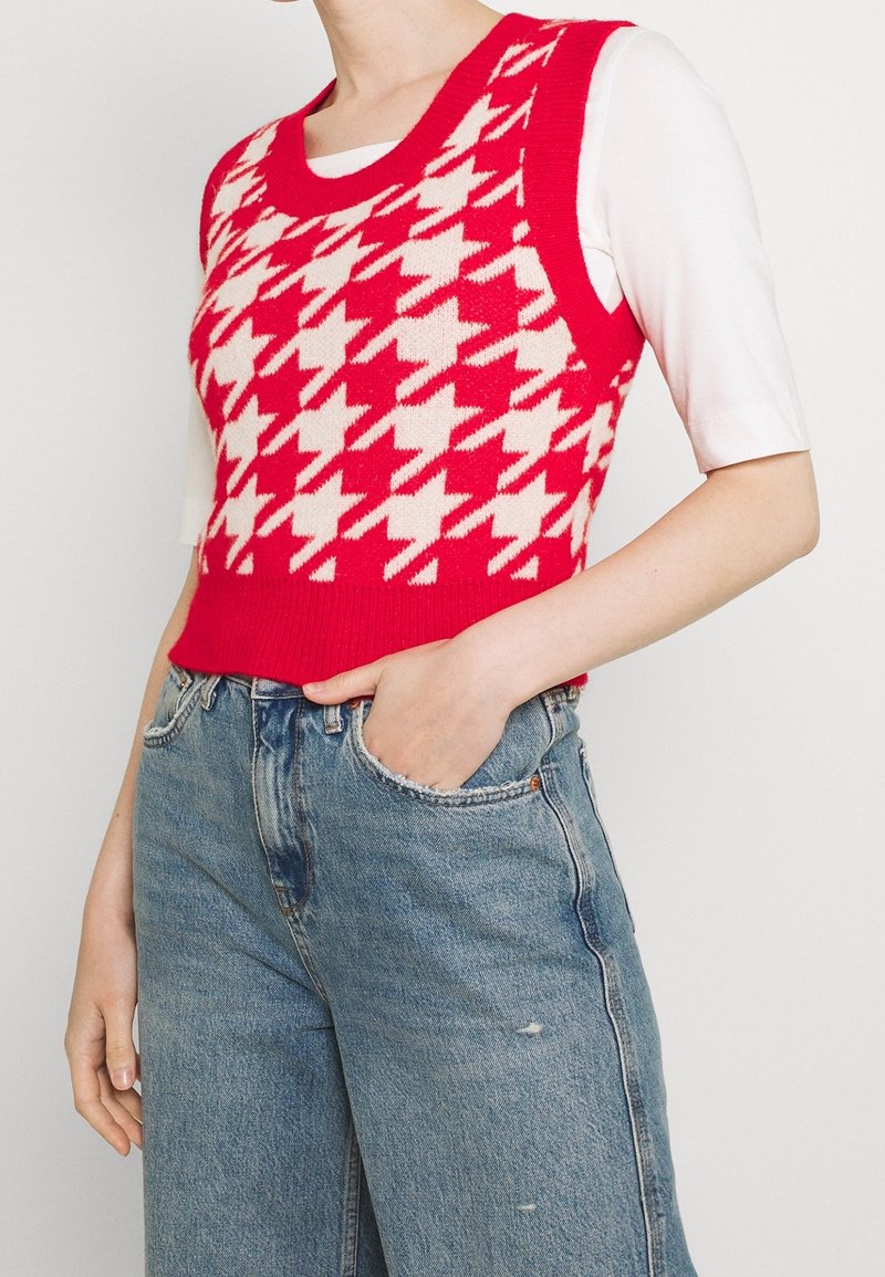 Czerwona i kremowa dzianinowa kamizelka w pepitkę z okrągłym dekoltem, noszona na białej dopasowanej bluzce i zestawiona z jasnoniebieskimi dżinsami z wysokim stanem.