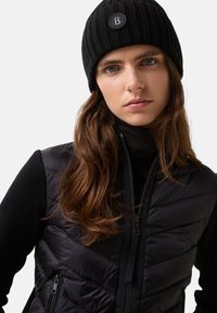 Czarna dzianinowa czapka typu beanie z prążkowaną teksturą i naszywką z logo; zestawiona z czarną puchową kamizelką z zapięciem na zamek i pikowanym wzorem.