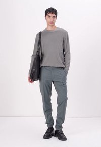 Grijze lange mouw shirt, lichtgrijze broek met subtiele textuur, en zwarte schoeisel. Model draagt een zwarte schoudertas. Minimalistisch ontwerp.