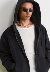 Corta-vento preto com fecho, com acabamento texturizado e bolsos, usado sobre uma t-shirt de alças brancas com textura de côr rib. Os acessórios incluem um colar de contas coloridas e óculos de sol ovais.