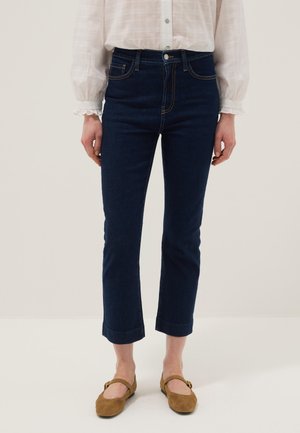 Vrouw met opgerolde donkerblauwe rechte jeans, witte blouse met lange mouwen en gerimpelde manchetten, en bruine Mary Jane ballerina's staand.