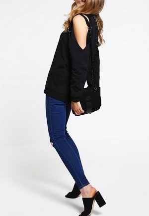 Musta avoselkäinen collegepaita, siniset kapeat farkut, joissa on kulunut polvi. Musta käsilaukku, jossa on ketjuhihna. Mustat korkeat mules-kengät. Yksinkertainen tausta.