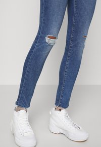 Valkoiset korkeakorkoiset tennarit, joissa on kuitukangas, yhdistettynä tiukkoihin sinisiin farkkuihin, joissa on kuluneita kohtia ja revittyjä helmoja.