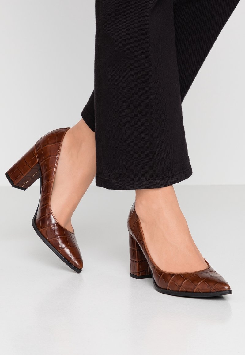 Femme portant des escarpins marron foncé à talon bloc imprimé crocodile associés à un pantalon court noir sur un fond clair.
