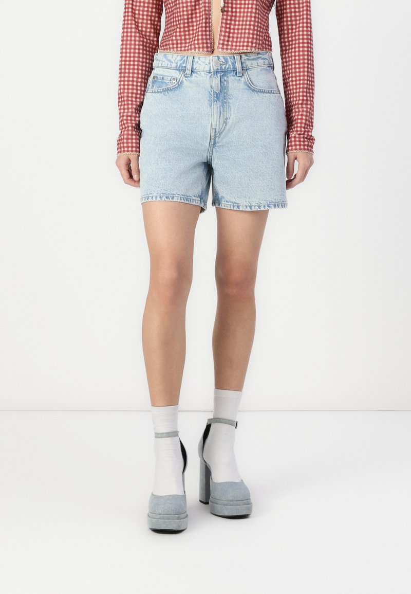Shorts en denim bleu clair avec un ourlet effiloché, dotés d'un design à cinq poches et d'une fermeture à bouton à l'avant, associés à des chaussures à plateforme grises et des chaussettes blanches.