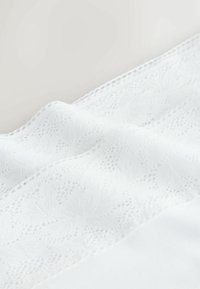 Tissu blanc doux avec des motifs géométriques complexes rappelant de la dentelle et un bord festonné, présenté en gros plan avec des plis délicats.