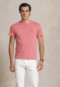 Jauns vīrietis, tērpies rozā īsa piedurkņu t-kreklā un baltās biksēs, stāv ar rokām atslābinātā pozā pret neitrālu fona.