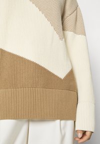 Hřejivý pletený svetr s béžovým, krémovým a bílým vzorem ve tvaru V, s manžetami na rukávech a žebrovaným lemem, nošený s bílými kalhotami.