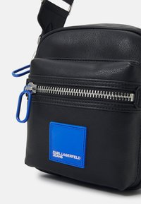 Crna kožna torbica preko tijela s metalnim zatvaračem, plavom oznakom i podesivim remenom. Kompaktni dizajn s teksturiranom površinom.