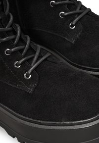 Botas de ante negras con cordones redondos, ojales de metal y una suela negra gruesa y texturizada. La parte superior presenta un acabado suave y detalles sutiles.