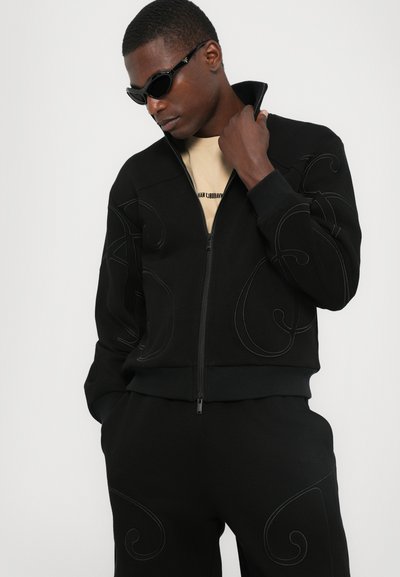 Sweatshirt noir zippé avec un pantalon assorti, présentant des motifs en relief en spirale. Texture lisse, coupe décontractée et design à col haut.