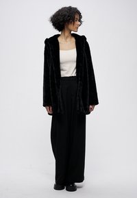 Melnas mākslīgās kažokādas mētelis ar kapuci, atvērta priekšpuse un gariem piedurknēm. Kombinēts ar krēmkrāsas topu un melnām biksēm ar platām kāju daļām, ar gludu tekstūru.