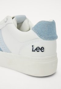 Zapatilla blanca con acentos en azul claro, detalles perforados y un talón de tela vaquera. Logo "Lee" bordado en negro en el lateral. Suela de goma.