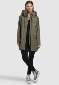 Parka vert olive avec une capuche, poches avant, fermeture éclair et boutons-pression. Porté par-dessus des leggings noirs et des baskets montantes vertes.