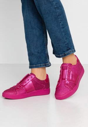Blue jeans worn with shiny pink sneakers featuring star decorations and a wide strap across the top, standing on a light floor.