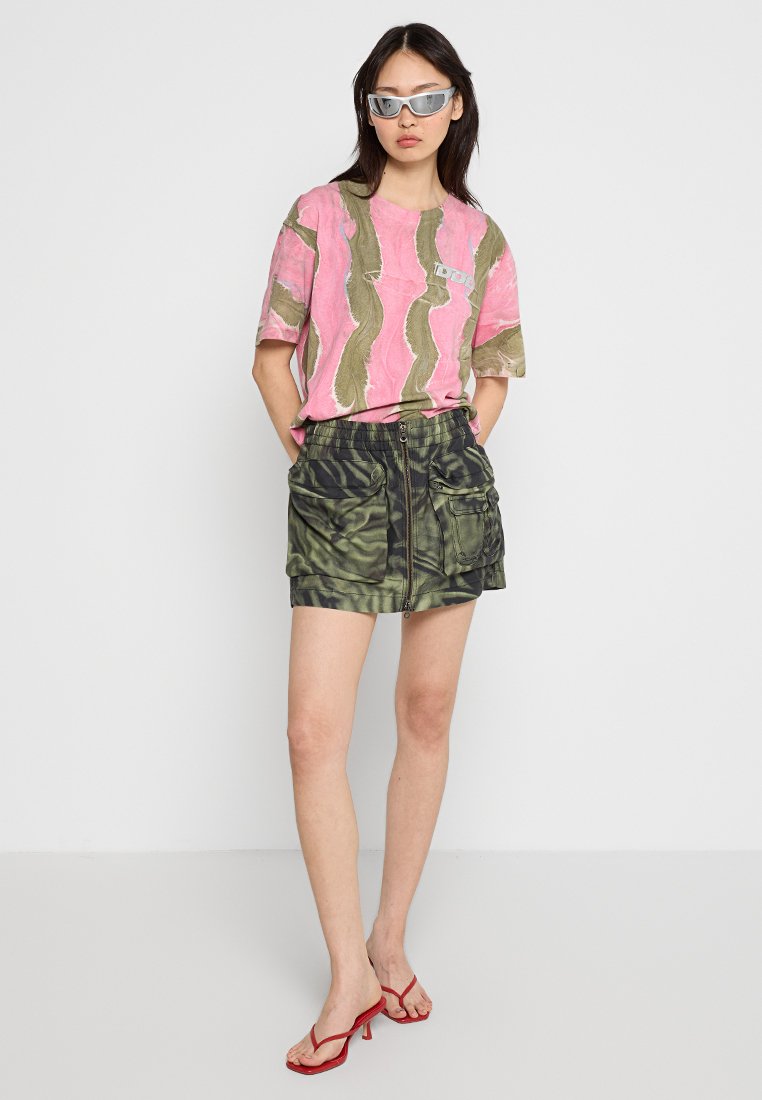 Donna che indossa una maglietta con stampa astratta rosa e oliva, una mini gonna cargo verde mimetica con zip, occhiali da sole argentati e sandali alti rossi.