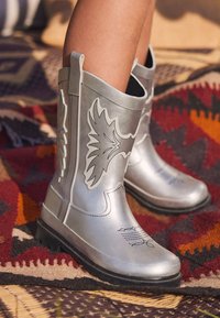 Sølvfarvede gummistøvler med sorte vingemønstre, flade såler og ankelhøjde. Tekstureret overdel med dekorative syningsdetaljer.