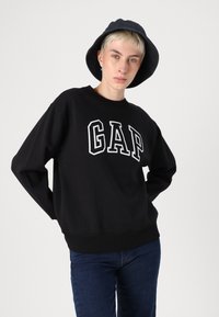 Juoda džemperis su baltu "GAP" logotipu, laisvas kirpimas, apvalus iškirpimas, elastingi rankogaliai ir apačia. Dėvimas su tamsiomis džinsinėmis kelnėmis ir juodu kibiru.