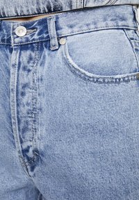 Vaqueros de mezclilla azul claro con un acabado ligeramente desgastado, que cuentan con un bolsillo frontal, cierre de botón metálico y detalles de costura visibles.