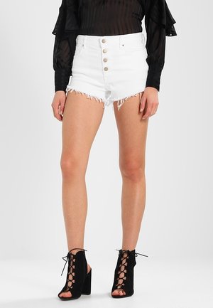 Shorts en denim blanc avec des bords effilochés, présentant une taille haute et une fermeture à cinq boutons à l'avant. Portés avec un haut noir transparent à manches longues et des escarpins noirs à lacets.