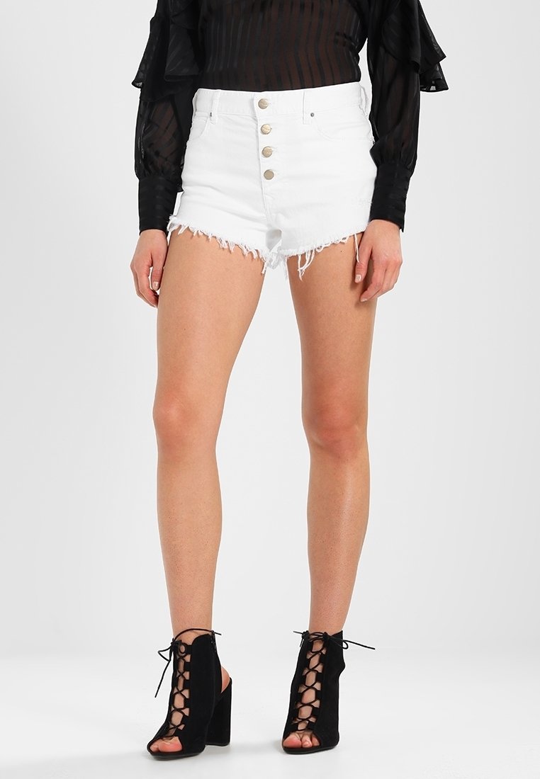 Shorts de mezclilla blanca con dobladillos deshilachados, que cuentan con una cintura alta y un cierre frontal de cinco botones. Combinados con una blusa negra de manga larga transparente y tacones negros con cordones.