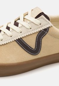 Baskets en daim marron clair avec des lacets crème, des accents en cuir brun foncé, et un logo ondulé distinctif sur le côté avec des coutures blanches.