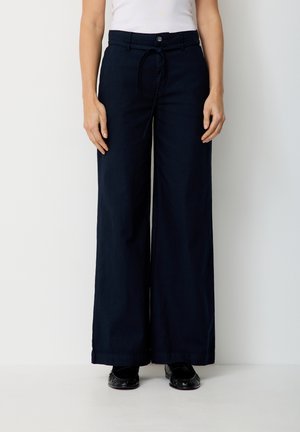 Frau trägt schwarze, hochtaillierte, weit geschnittene Hosen mit Knöpfen vorne und Tunnelzug, kombiniert mit schwarzen Loafers und weißem Oberteil.