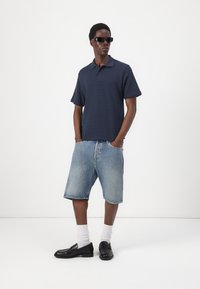 Mornarsko teksturirana polo majica s kratkimi rokavi, v kombinaciji s svetlo modrimi jeans kratkimi hlačami. Črne loafers čevlje in bele crew nogavice dopolnjujejo obleko.