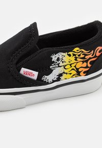 Svart kanvas slip-on sko med en lejon grafik med orange lågor, vit gummisula och röd Vans-tagg på sidan.