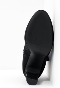 Bottes en daim noir vues de côté de la semelle, montrant une semelle lisse avec une légère rainure et un talon bloc sur un fond blanc.