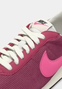 Růžové tenisky Nike mají texturovaný svršek připomínající manšestr, bílé šněrování a výrazné růžové logo. Design je sportovní a moderní.
