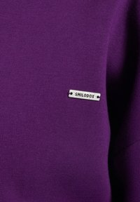 Tessuto viola con una finitura testurizzata, completato da un piccolo tag argentato con "SMILODOX" impresso.