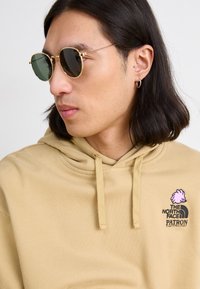 Felpa beige con cappuccio e coulisse, caratterizzata da un design ricamato rosa e un logo nero sul petto. Il modello indossa occhiali da sole rotondi color oro.